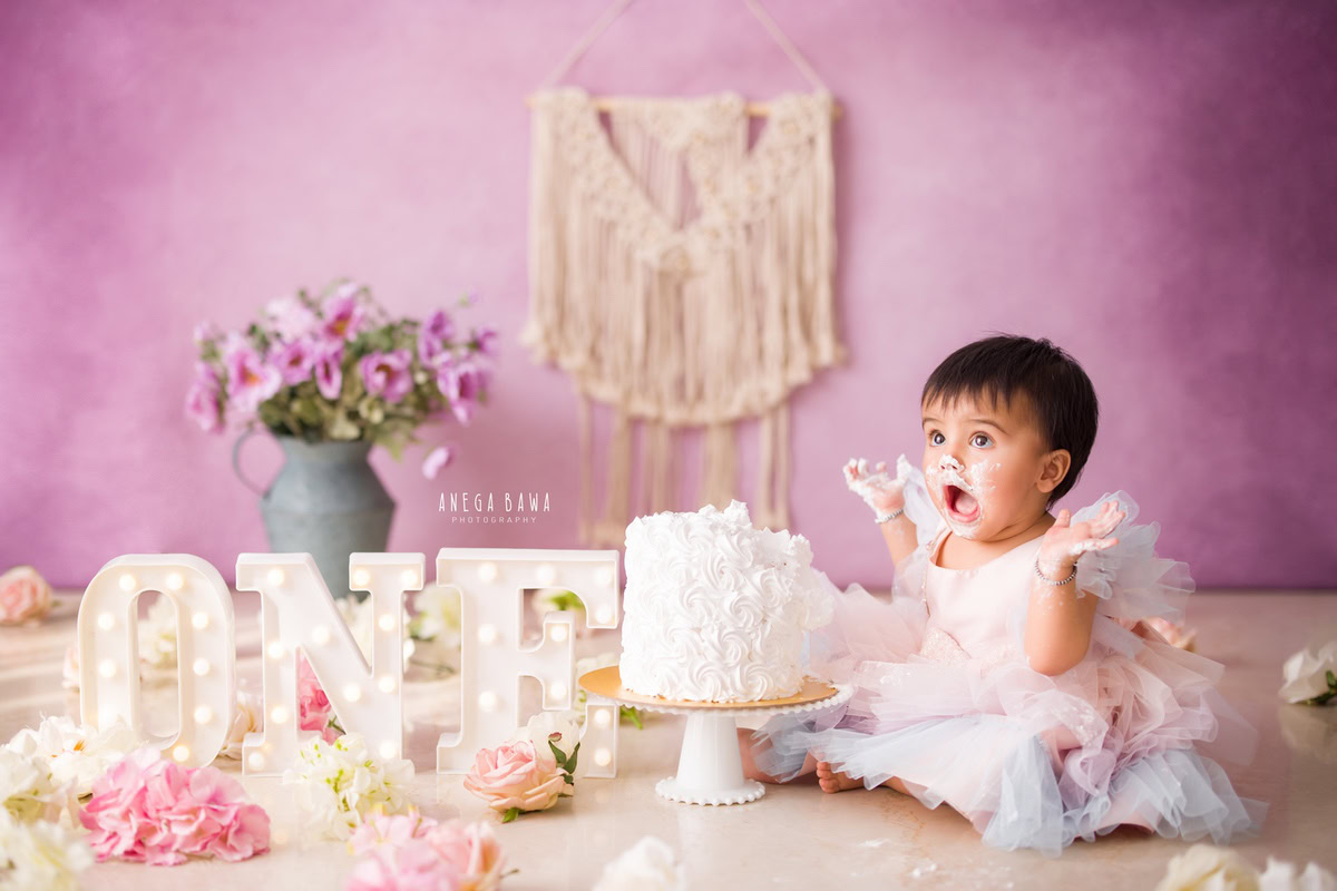 1 year old girl with lavender backdrop, vibrant Cake Smash, pink and white flowers, and a green flower vase, first birthday photography in Delhi, Gurgaon. Captured by Anega Bawa family photographer Gurgaon (Delhi NCR)