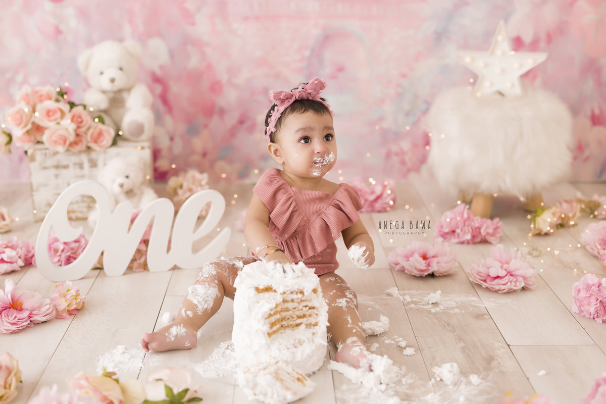 1 year old girl with a pink backdrop, joyful Cake Smash, surrounded by cuddly teddy bears and beautiful flowers, first birthday photoshoot in Delhi, Gurgaon. Captured by Anega Bawa family photographer Gurgaon (Delhi NCR)