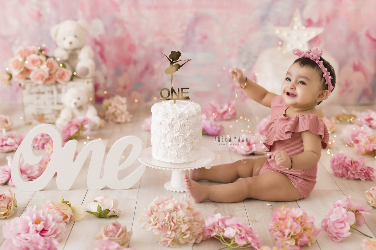 1 year old girl with a pink backdrop, playful Cake Smash, surrounded by teddy bears and peach-pink flowers on the floor, first birthday photography in Delhi, Gurgaon. Captured by Anega Bawa family photographer Gurgaon (Delhi NCR)