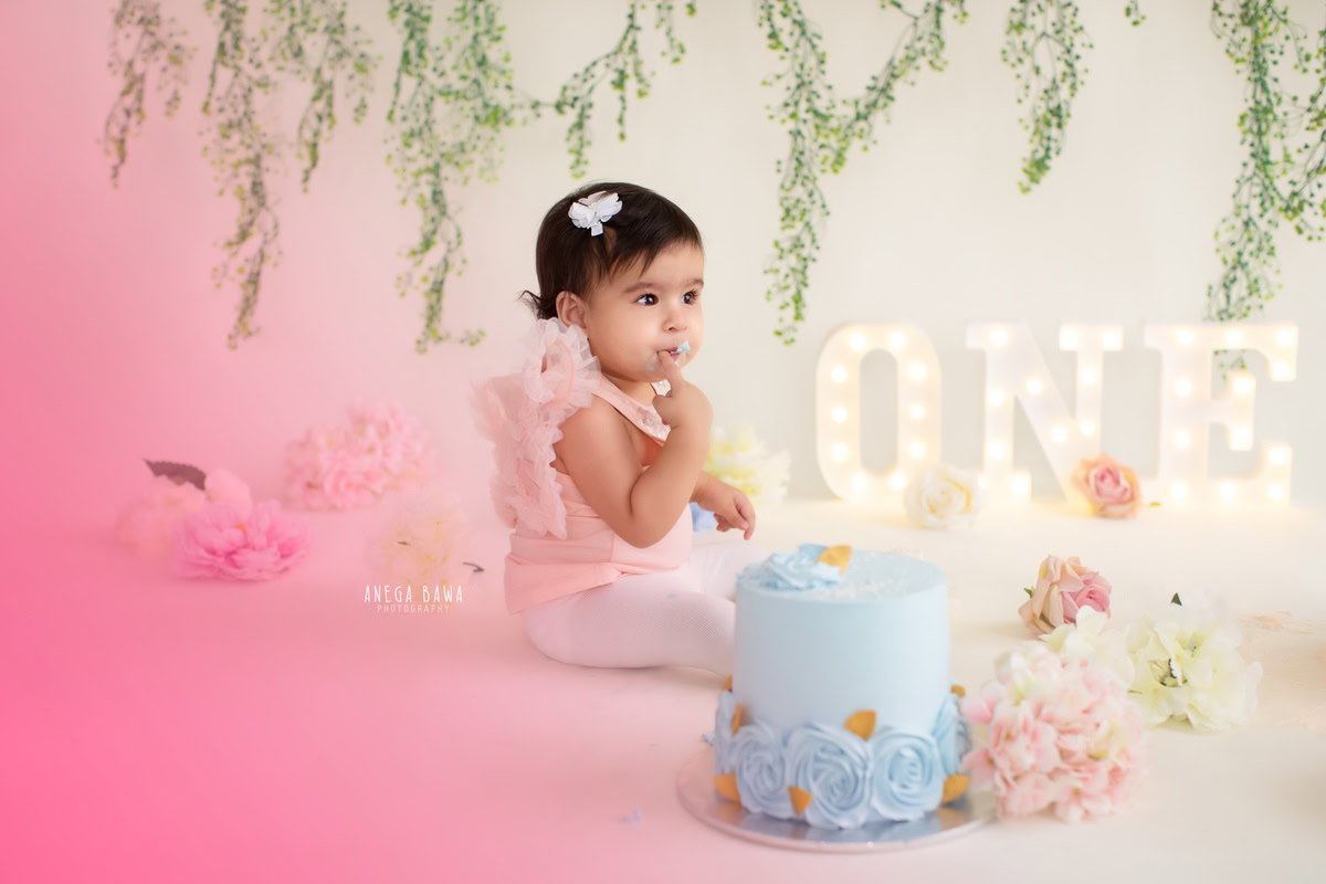 1 year old girl with a pink backdrop, leafy fringe on the wall, and vibrant Cake Smash, first birthday photoshoot in Delhi, Gurgaon. Captured by Anega Bawa family photographer Gurgaon (Delhi NCR)