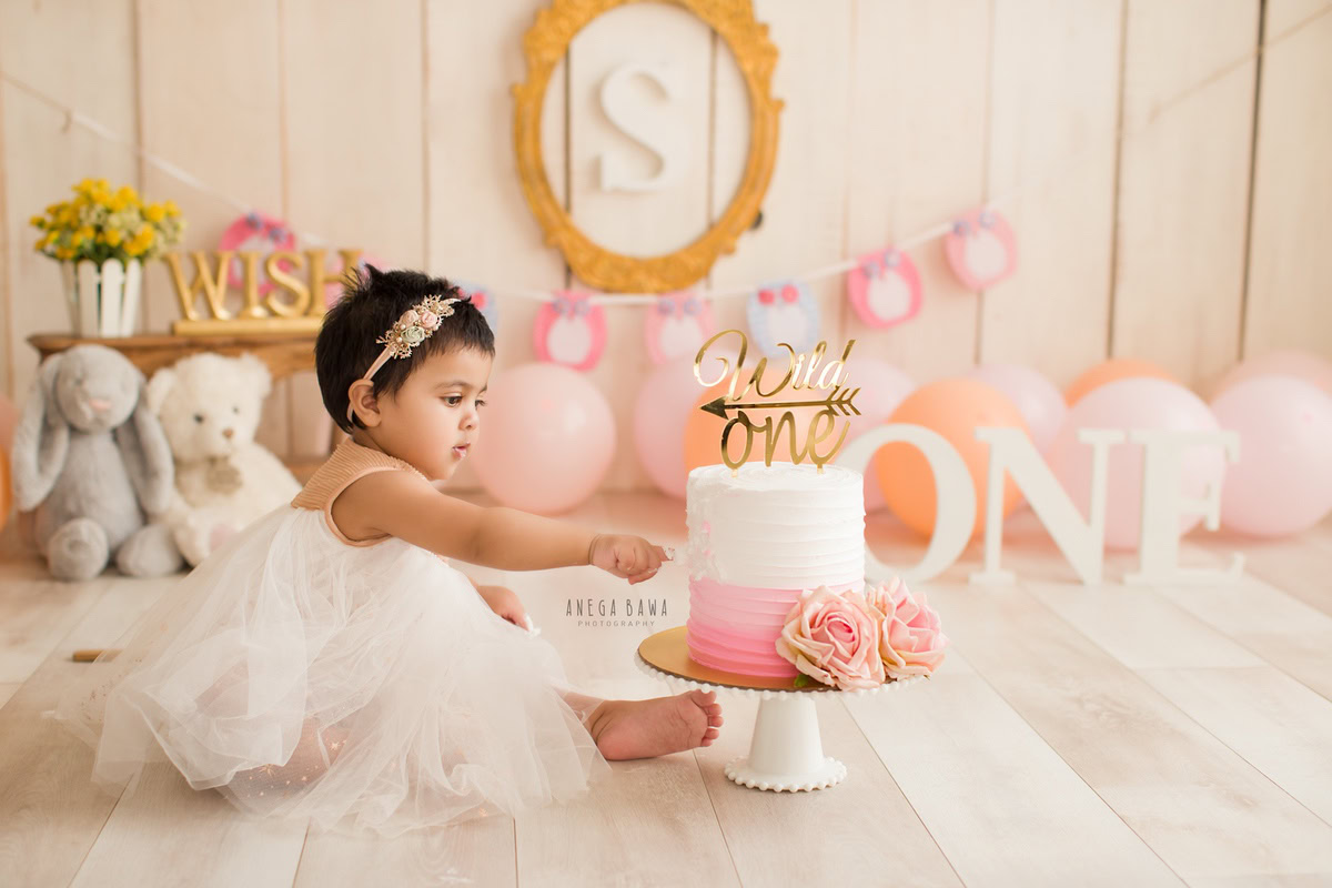 1 year old girl with pink balloons, teddy bears, and a golden frame on the wall, set against a beige backdrop. Celebrating her first birthday. Cake Smash photo shoot in Delhi, Gurgaon by Anega Bawa Family Photographer Gurgaon (Delhi NCR).