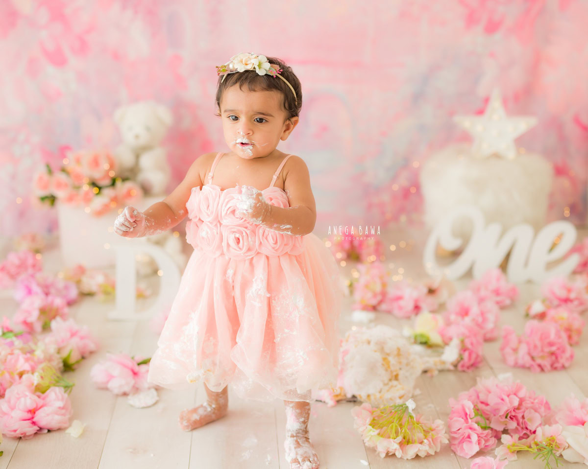 1 year old girl in a pink dress with cake smash on her face against a pink backdrop with flowers on the floor. Captured during a charming first birthday photography session in Delhi Gurgaon by the renowned family photographer Anega Bawa.