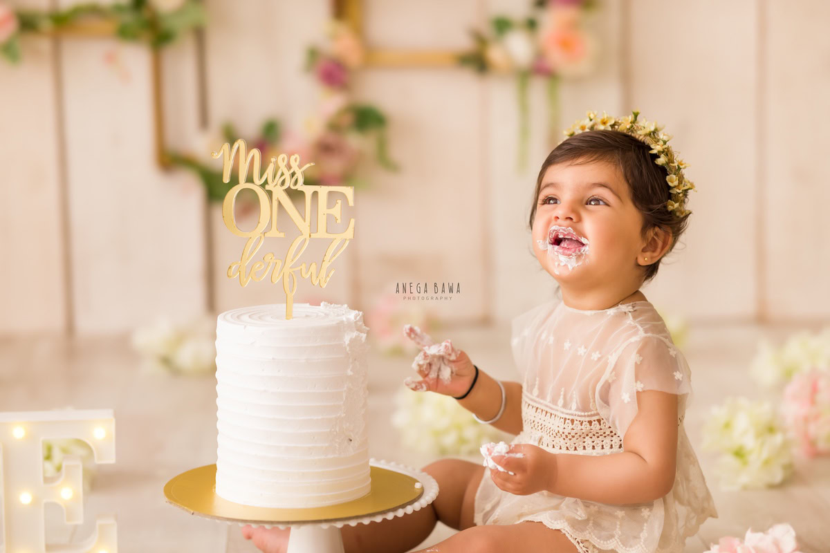 1 year old girl playing with Cake Smash, beige backdrop, floral frame on the wall, and delicate white flowers, first birthday photoshoot in Delhi, Gurgaon. Captured by Anega Bawa family photographer Gurgaon (Delhi NCR)