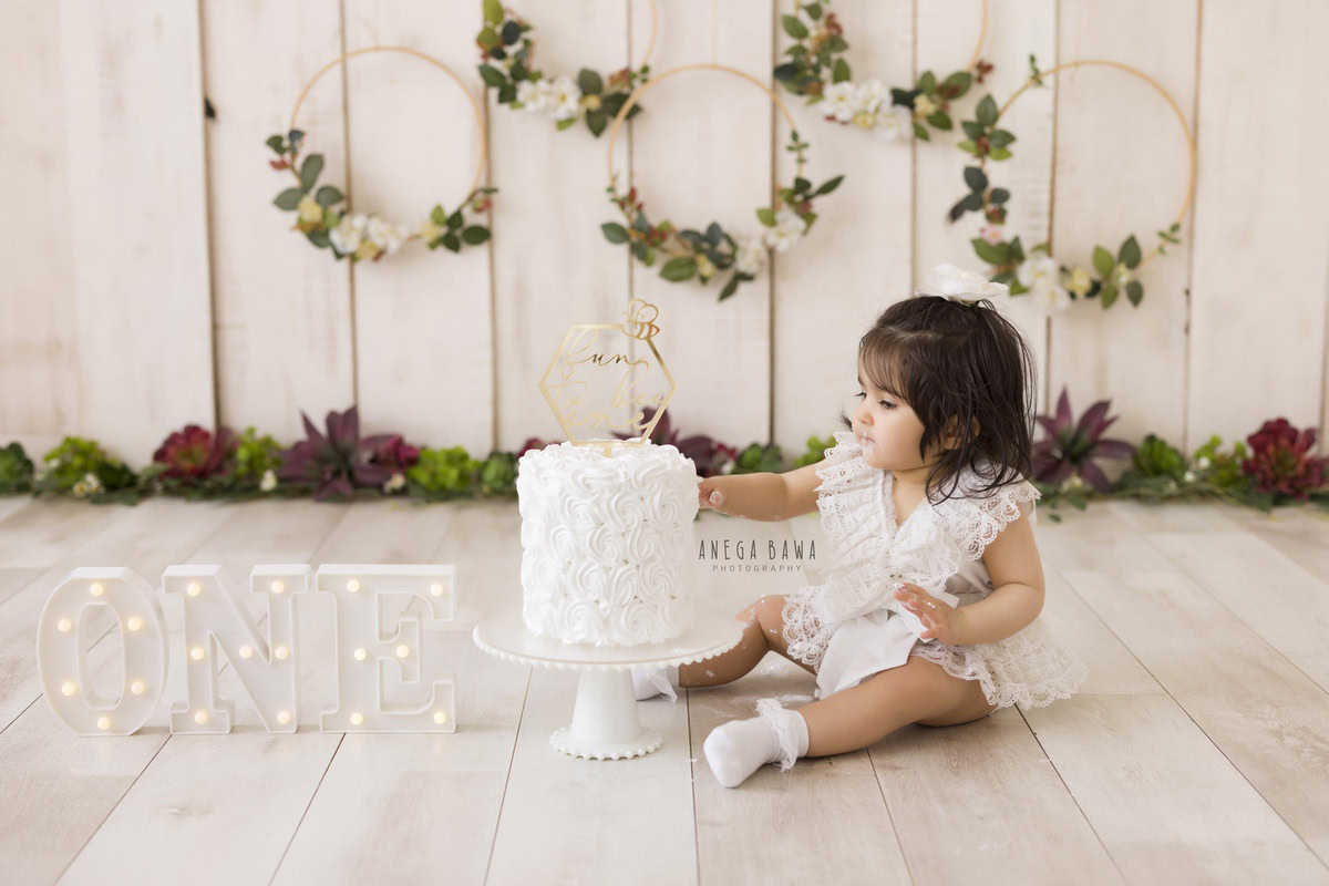 1year old girl playing with a cake smash setup against a beige backdrop, featuring a round wooden floral frame on the wall, captured by Anega Bawa during a Cake Smash photoshoot in Delhi, Gurgaon