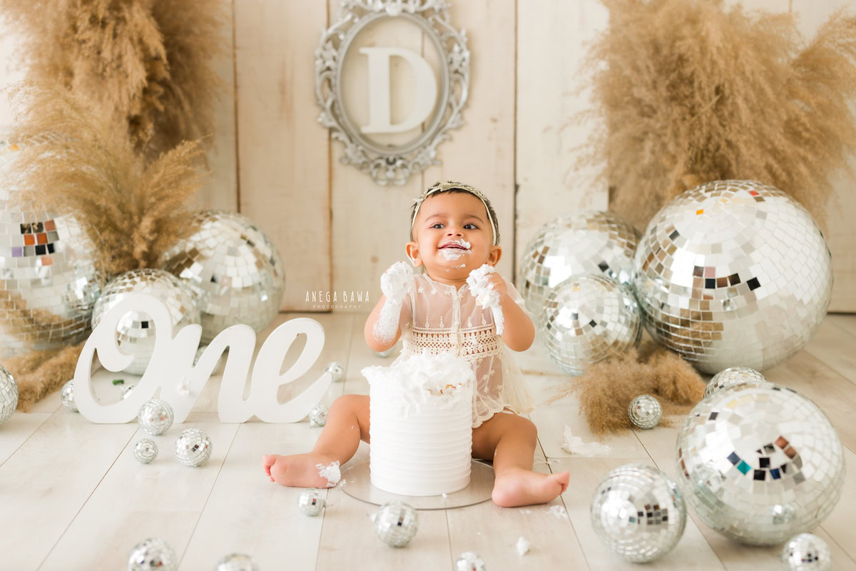 1 year old girl playing with cake smash surrounded by discoballs and bushes against a beige backdrop. Captured during a fun first birthday photography session in Delhi Gurgaon by the renowned family photographer Anega Bawa.