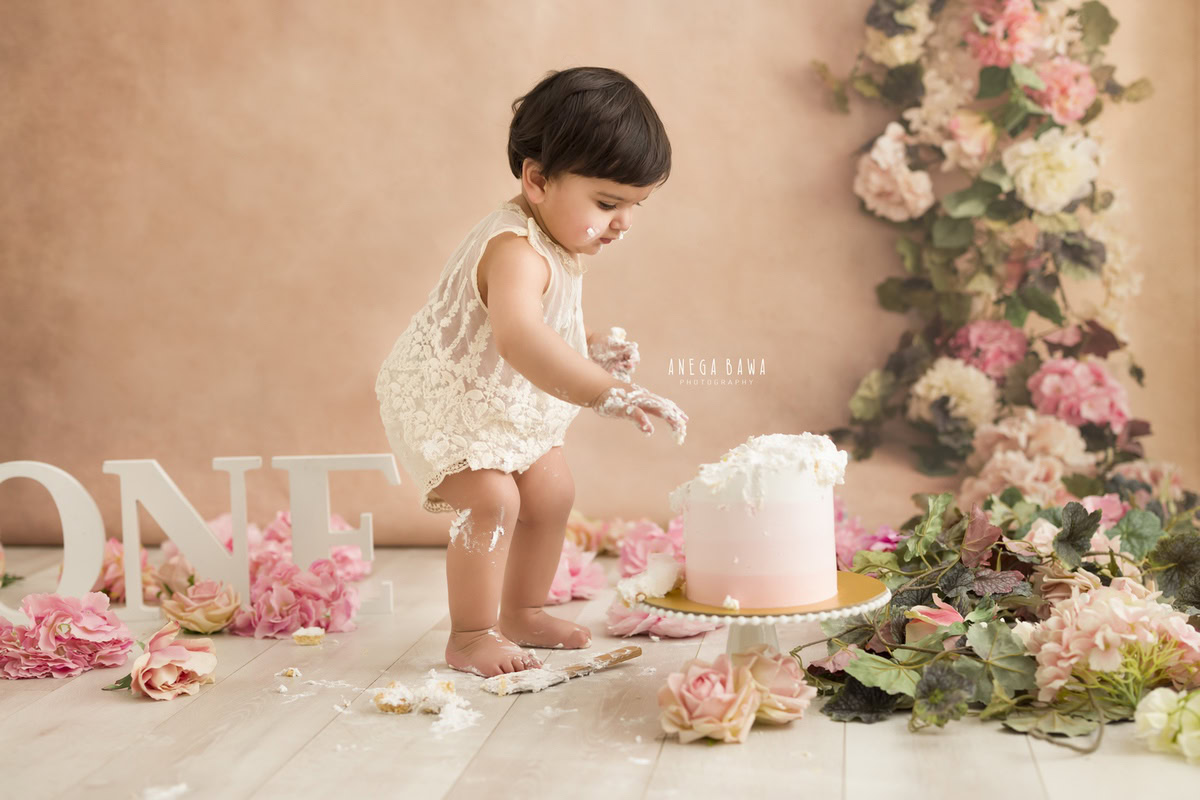 1 year old girl playing with cake smash against a peach backdrop with pink flowers on the floor. Captured during a delightful first birthday photography session in Delhi Gurgaon by the renowned family photographer Anega Bawa.