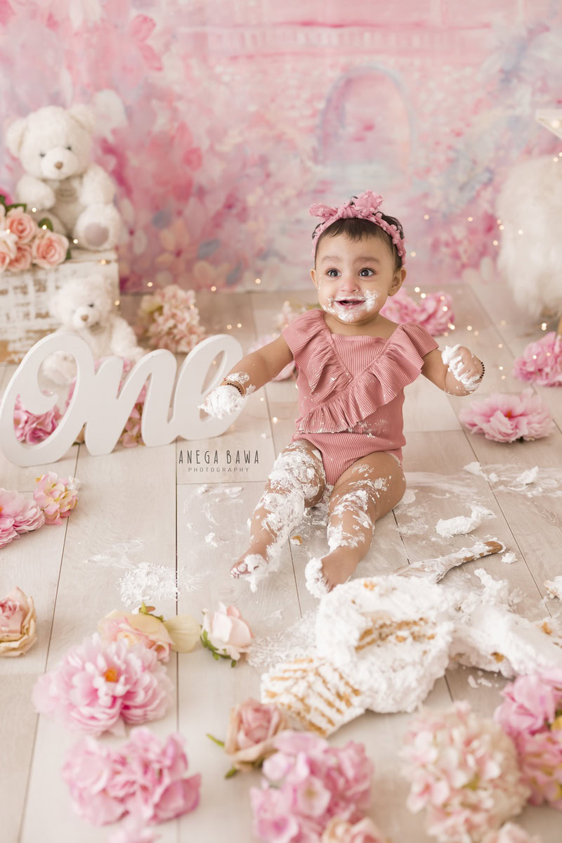 1year old girl playing with a cake smash setup against a pink backdrop with flowers on the floor, captured by Anega Bawa during a Cake Smash photoshoot in Delhi, Gurgaon
