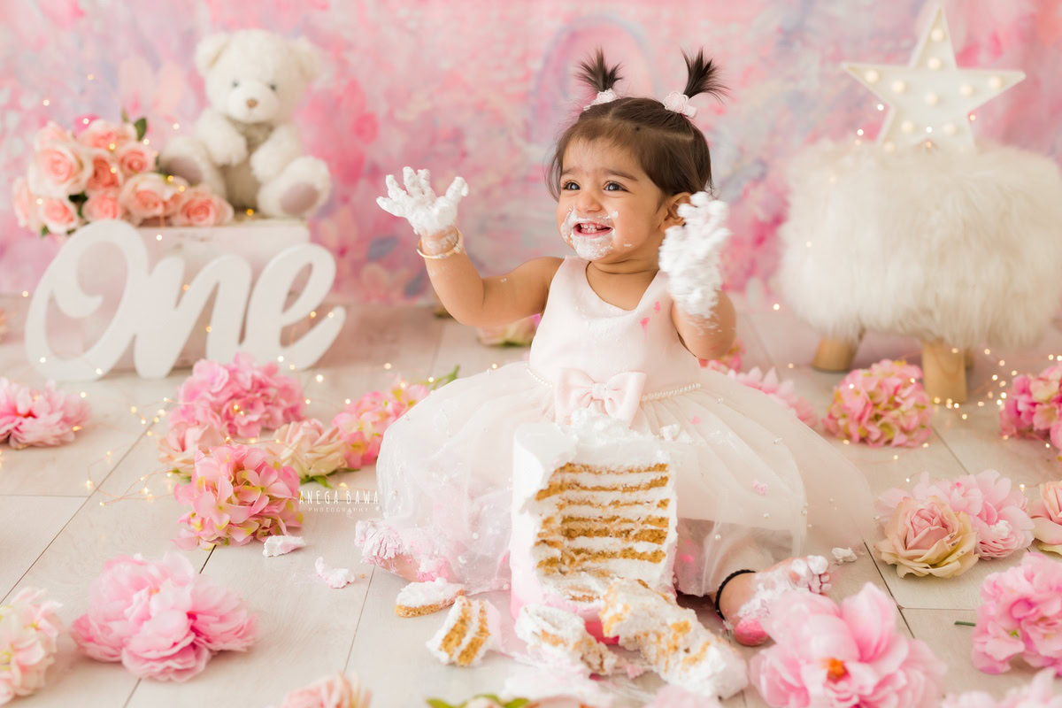 1 year old girl playing with cake smash against a pink backdrop with teddybears and flowers. Captured during a charming first birthday photoshoot in Delhi Gurgaon by the renowned family photographer Anega Bawa.