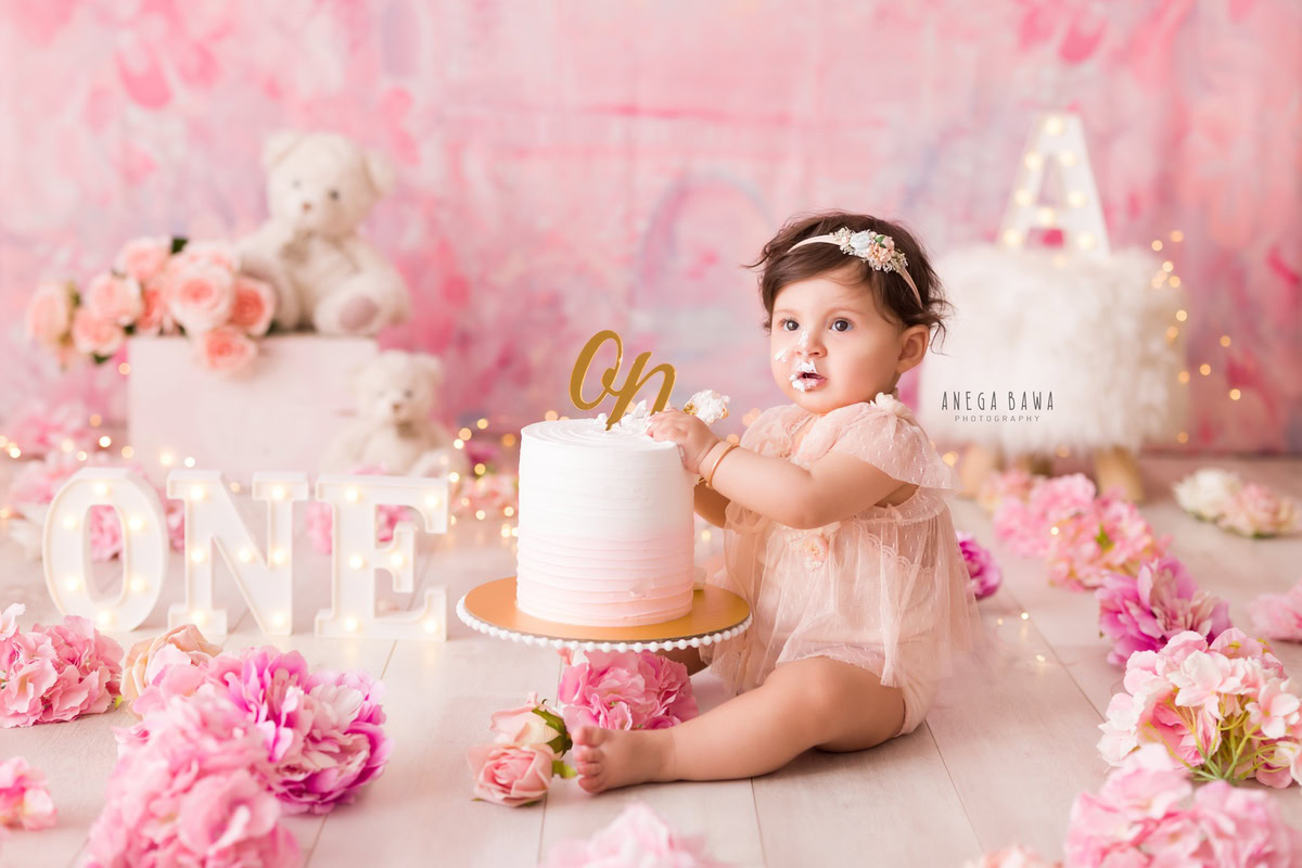 1 year old girl playing with a cake smash setup, featuring a pink backdrop, teddy bears, pink and peach flowers, and a cute headband. Celebrating her first birthday. Cake Smash photo shoot in Delhi, Gurgaon by Anega Bawa Family Photographer Gurgaon (Delhi NCR).