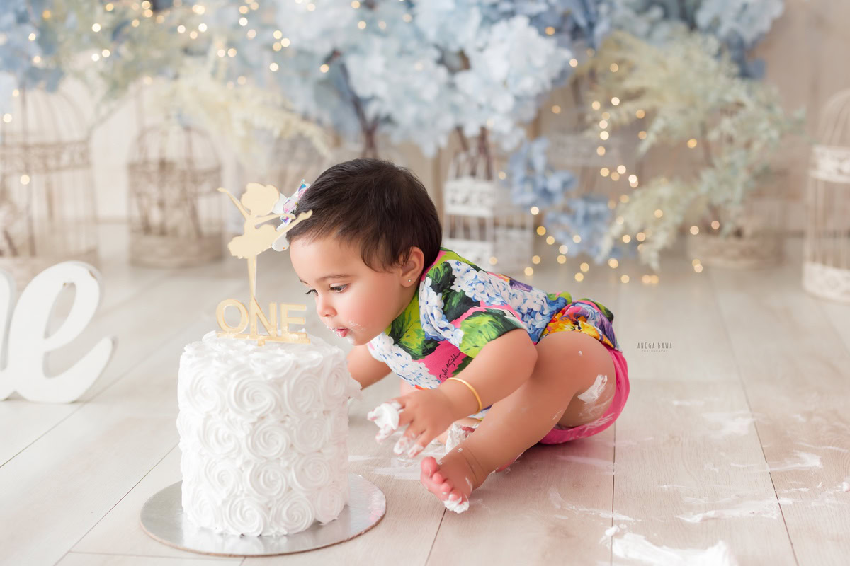 1 year old girl playing with Cake Smash, surrounded by whimsical white castles and twinkling fairy lights, first birthday photoshoot in Delhi, Gurgaon. Captured by Anega Bawa family photographer Gurgaon (Delhi NCR)