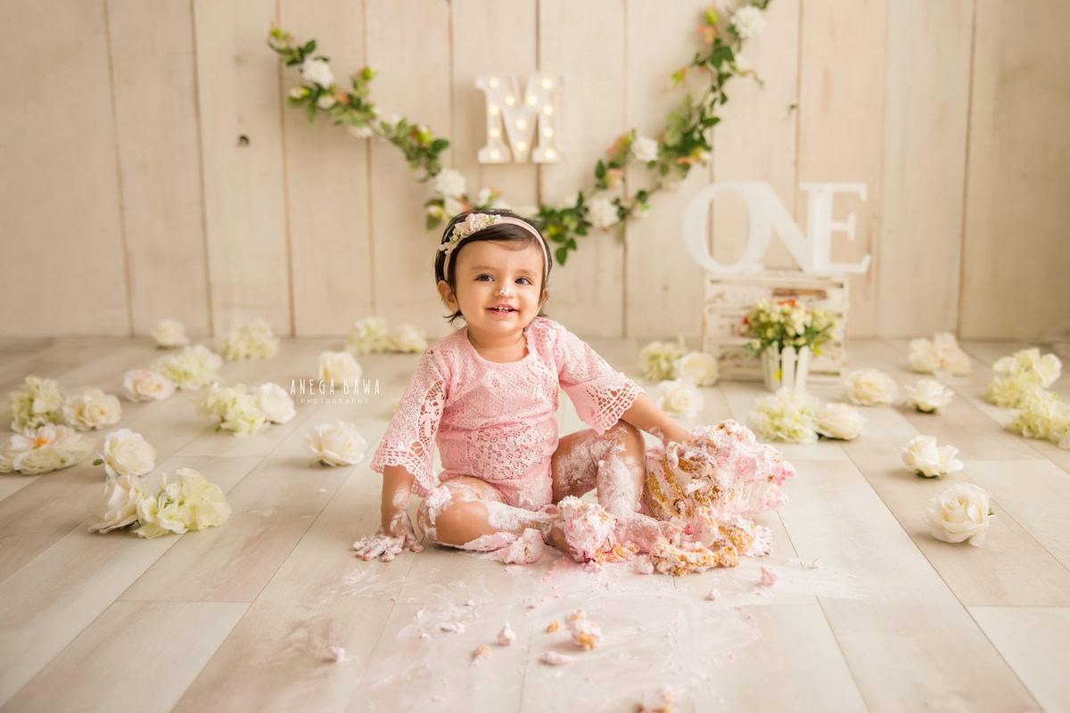 1 year old girl playing with Cake Smash, surrounded by white flowers, a beige backdrop, and a floral fringe on the wall, first birthday photo shoot in Delhi, Gurgaon. Captured by Anega Bawa family photographer Gurgaon (Delhi NCR)
