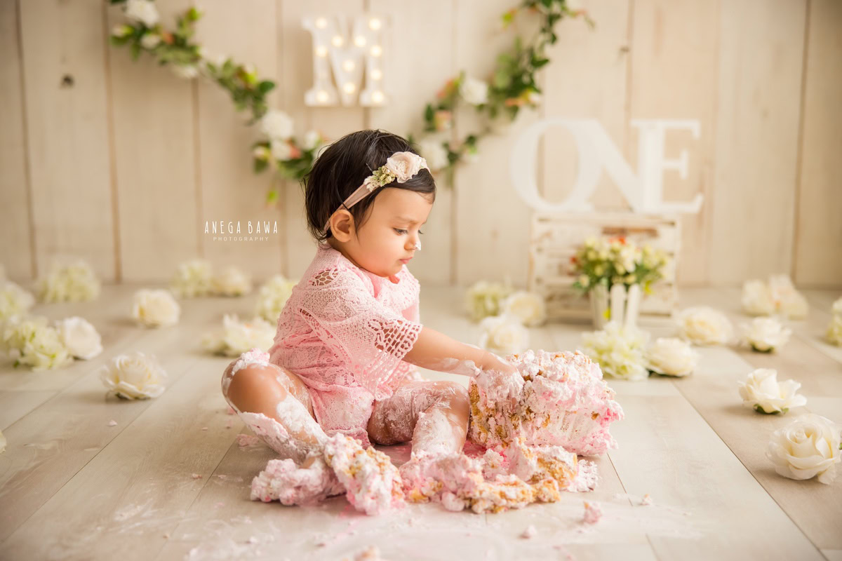 1 year old girl playing with cake smash surrounded by white flowers and a floral fringe on the wall. Captured during a delightful first birthday photography session in Delhi Gurgaon by the renowned family photographer Anega Bawa.