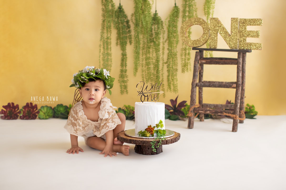 1 year old girl posing with a cake smash setup, featuring leafy fringes on the wall, a yellow backdrop, and a golden frame on a wooden stool. Celebrating her first birthday. Cake Smash photo shoot in Delhi, Gurgaon by Anega Bawa Family Photographer Gurgaon (Delhi NCR).