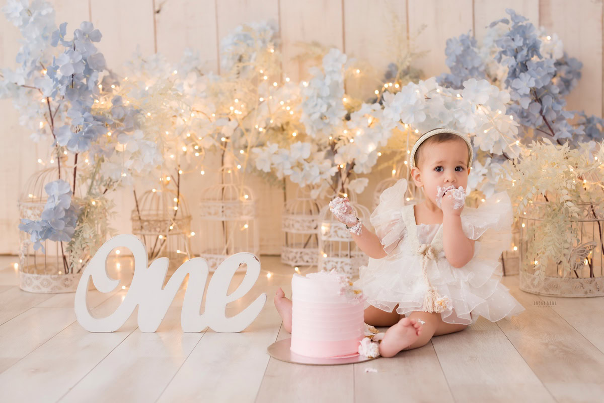 1 year old girl posing with Cake Smash, surrounded by blue and white flowers, a beige backdrop, and sparkling fairy lights, first birthday photoshoot in Delhi, Gurgaon. Captured by Anega Bawa family photographer Gurgaon (Delhi NCR)