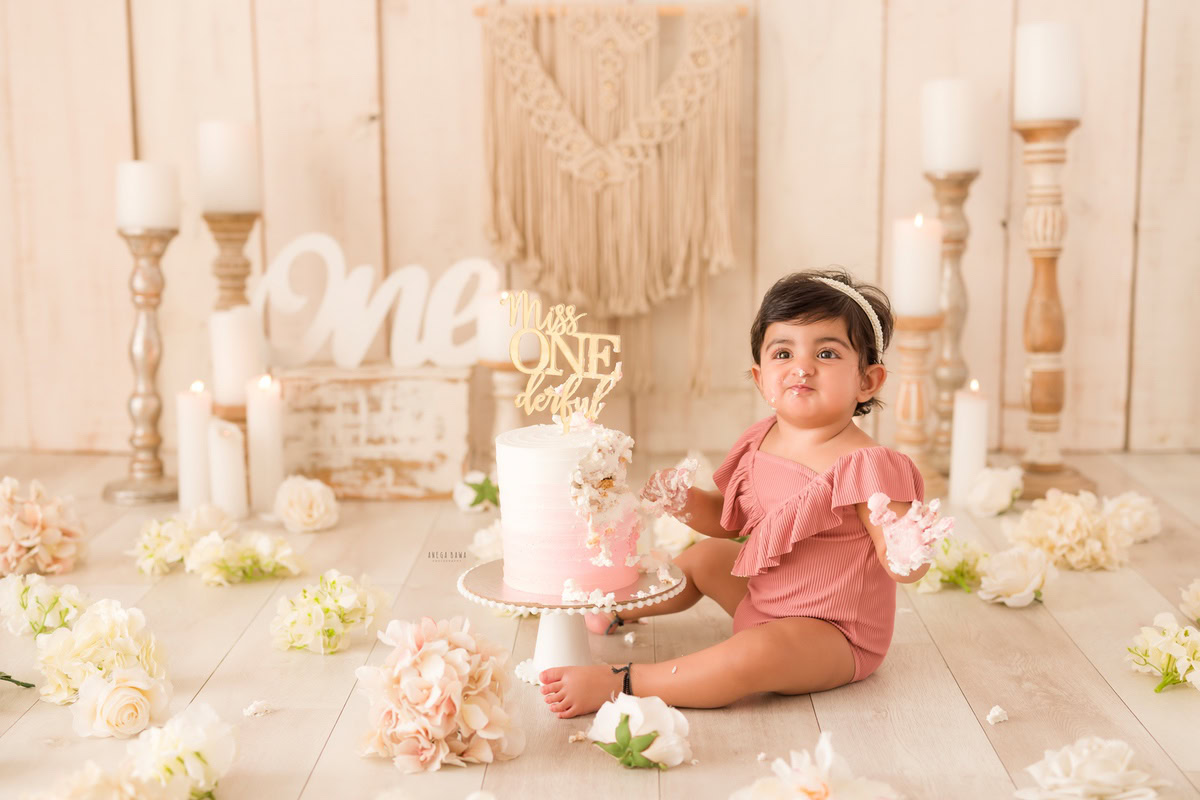 1year old girl posing with a cake smash setup and a candle stand against a beige backdrop, captured by Anega Bawa during a Cake Smash photoshoot in Delhi, Gurgaon