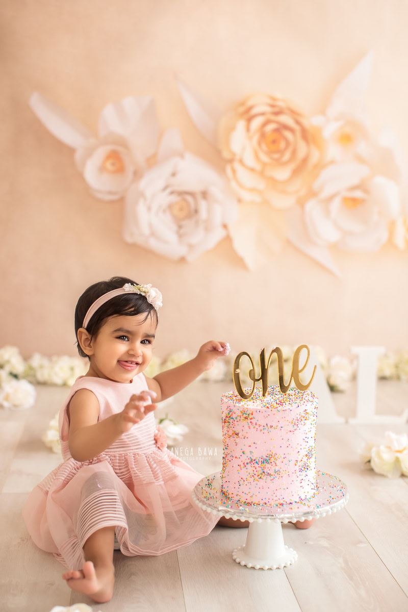 1 year old girl posing with a cake smash setup, featuring a floral arrangement and a beige backdrop. Celebrating her first birthday. Cake Smash photo shoot in Delhi, Gurgaon by Anega Bawa Family Photographer Gurgaon (Delhi NCR).