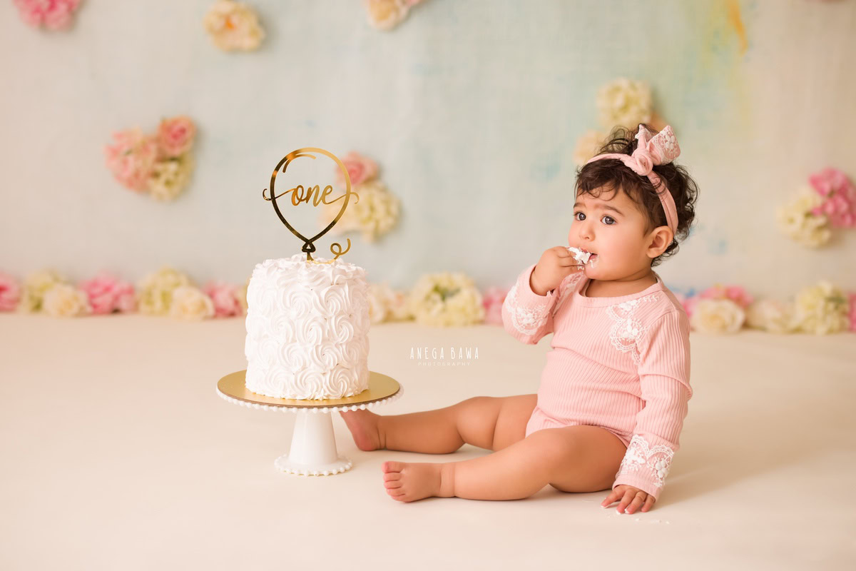 1 year old girl posing with a cake smash setup, featuring flowers, a cute headband, and a white backdrop. Celebrating her first birthday. Cake Smash photo shoot in Delhi, Gurgaon by Anega Bawa Family Photographer Gurgaon (Delhi NCR).