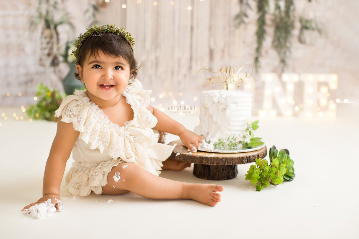 1 year old girl posing with a cake smash setup, featuring a leafy backdrop and fairy lights. Celebrating her first birthday. Cake Smash photo shoot in Delhi, Gurgaon by Anega Bawa Family Photographer Gurgaon (Delhi NCR).