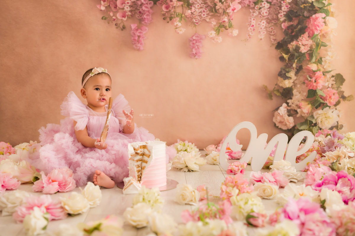 1year old girl posing with a cake smash setup against a peach backdrop, with flowers on the floor, captured by Anega Bawa during a Cake Smash photoshoot in Delhi, Gurgaon