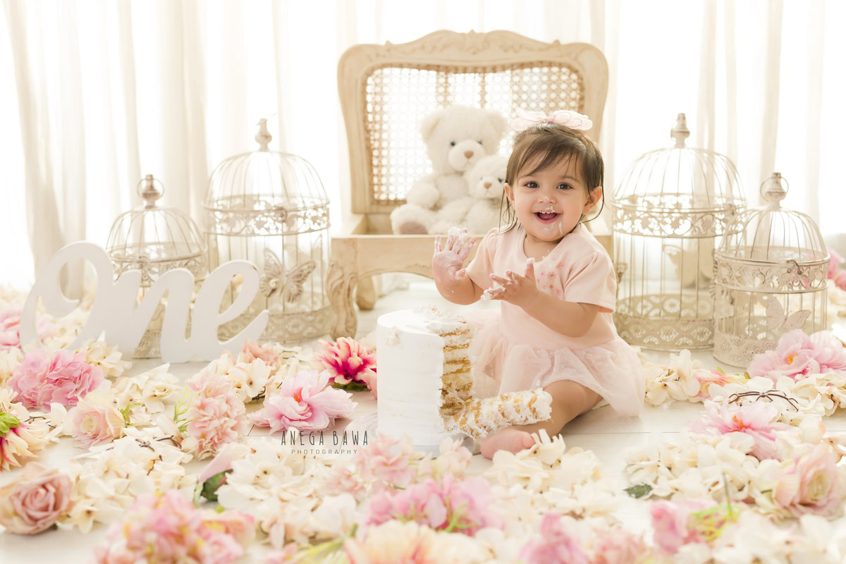 1 year old girl posing with Cake Smash, pink flowers on the floor, whimsical white castles, and a clean white backdrop, first birthday photo shoot in Delhi, Gurgaon. Captured by Anega Bawa family photographer Gurgaon (Delhi NCR)