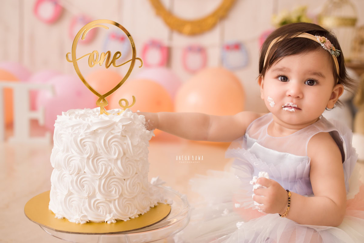 1 year old girl posing with Cake Smash, surrounded by pink and peach balloons against a beige backdrop, first birthday photography in Delhi, Gurgaon. Captured by Anega Bawa family photographer Gurgaon (Delhi NCR)