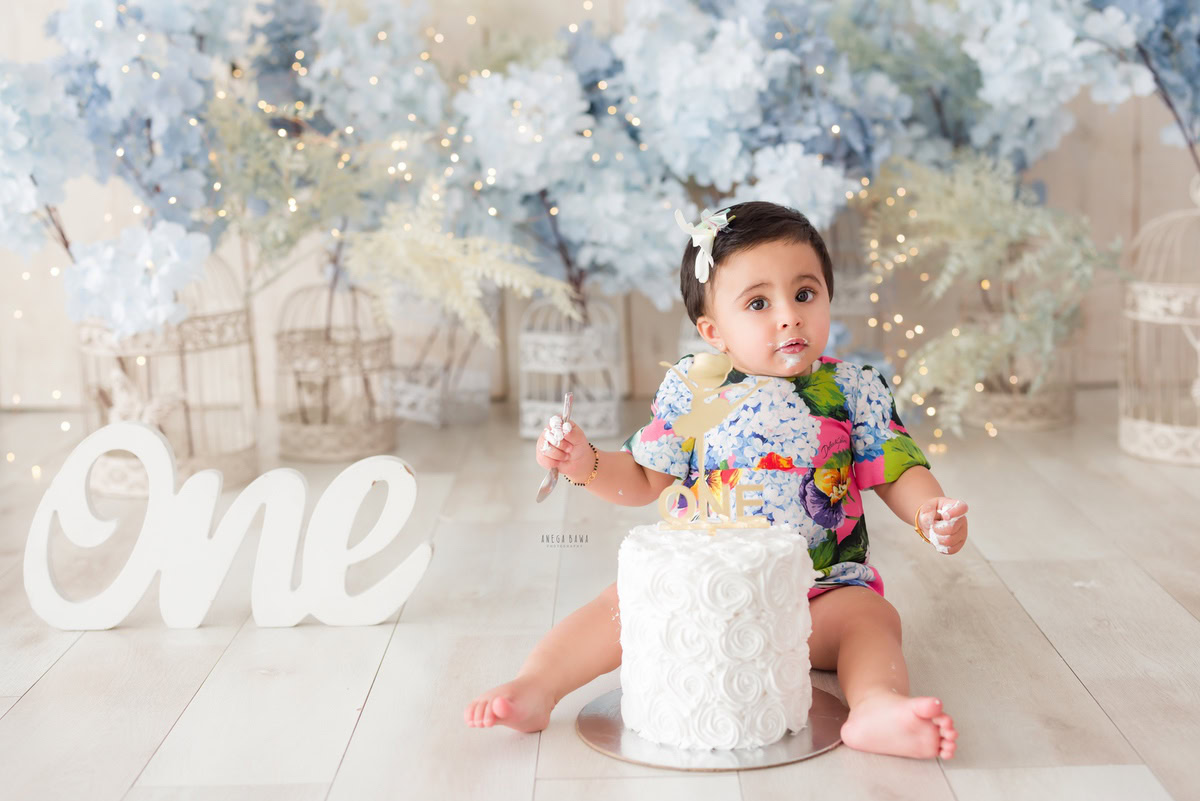 1year old girl posing with a cake smash setup against a white backdrop, featuring bunches of flowers, captured by Anega Bawa during a Cake Smash photoshoot in Delhi, Gurgaon