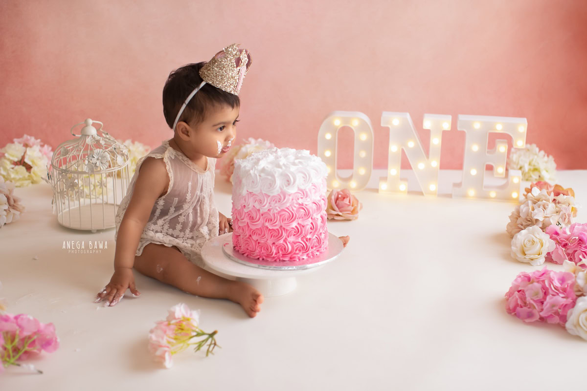 1 year old girl posing with a cake smash setup, featuring white castles, flowers, and a pink backdrop. Celebrating her first birthday. Cake Smash photo shoot in Delhi, Gurgaon by Anega Bawa Family Photographer Gurgaon (Delhi NCR).