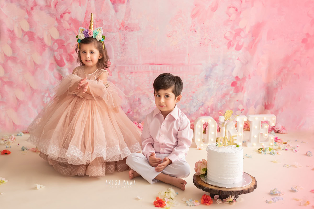1 year old girl and sibling with a cake smash setup, featuring a pink backdrop and flowers on the floor. Celebrating their first birthday. Cake Smash photo shoot in Delhi, Gurgaon by Anega Bawa Family Photographer Gurgaon (Delhi NCR).