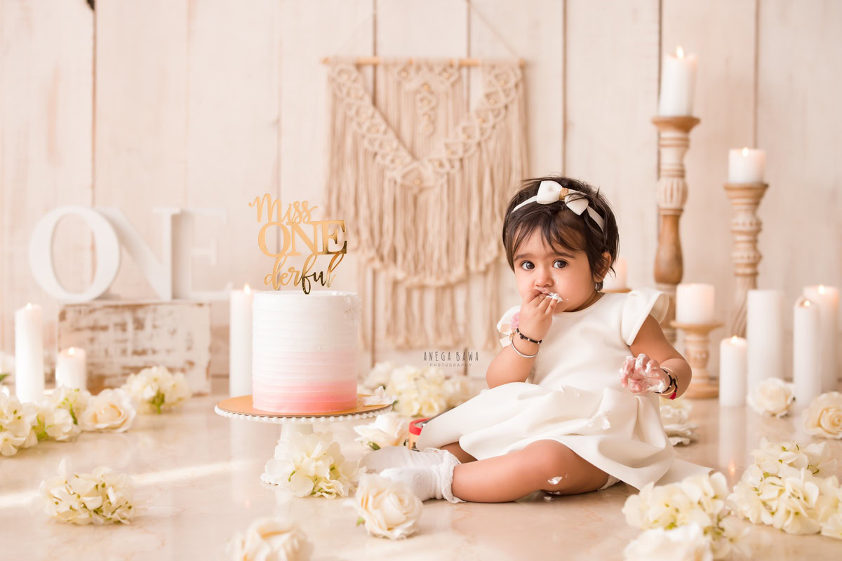 1 year old girl sitting on the floor with a cake smash setup, featuring a beige backdrop, a candle stand, and white flowers. Celebrating her first birthday. Cake Smash photo shoot in Delhi, Gurgaon by Anega Bawa Family Photographer Gurgaon (Delhi NCR).