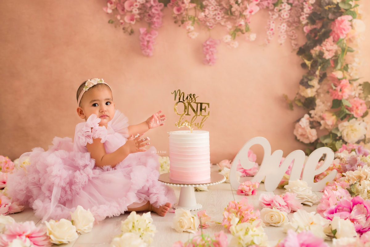 1year old girl sitting on the floor with a cake smash setup against a peach backdrop, featuring a floral fringe on the wall, captured by Anega Bawa during a Cake Smash photoshoot in Delhi, Gurgaon