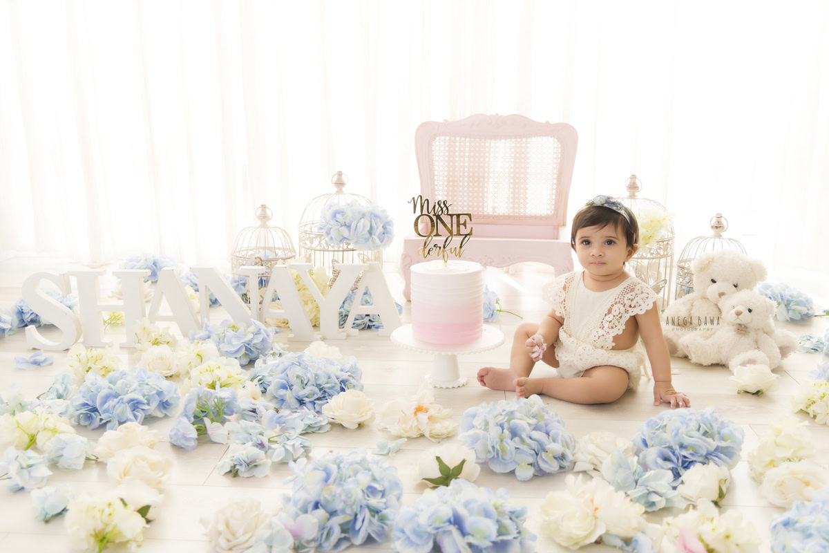 1year old girl sitting on the floor with a cake smash setup against a white backdrop, featuring blue and white flowers and a chair, captured by Anega Bawa during a Cake Smash photoshoot in Delhi, Gurgaon