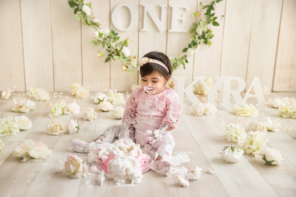 1 year old girl sitting on the floor with cake smash surrounded by white flowers and a floral fringe on the wall against a beige backdrop. Captured during a delightful first birthday photo shoot in Delhi Gurgaon by the renowned family photographer Anega Bawa.