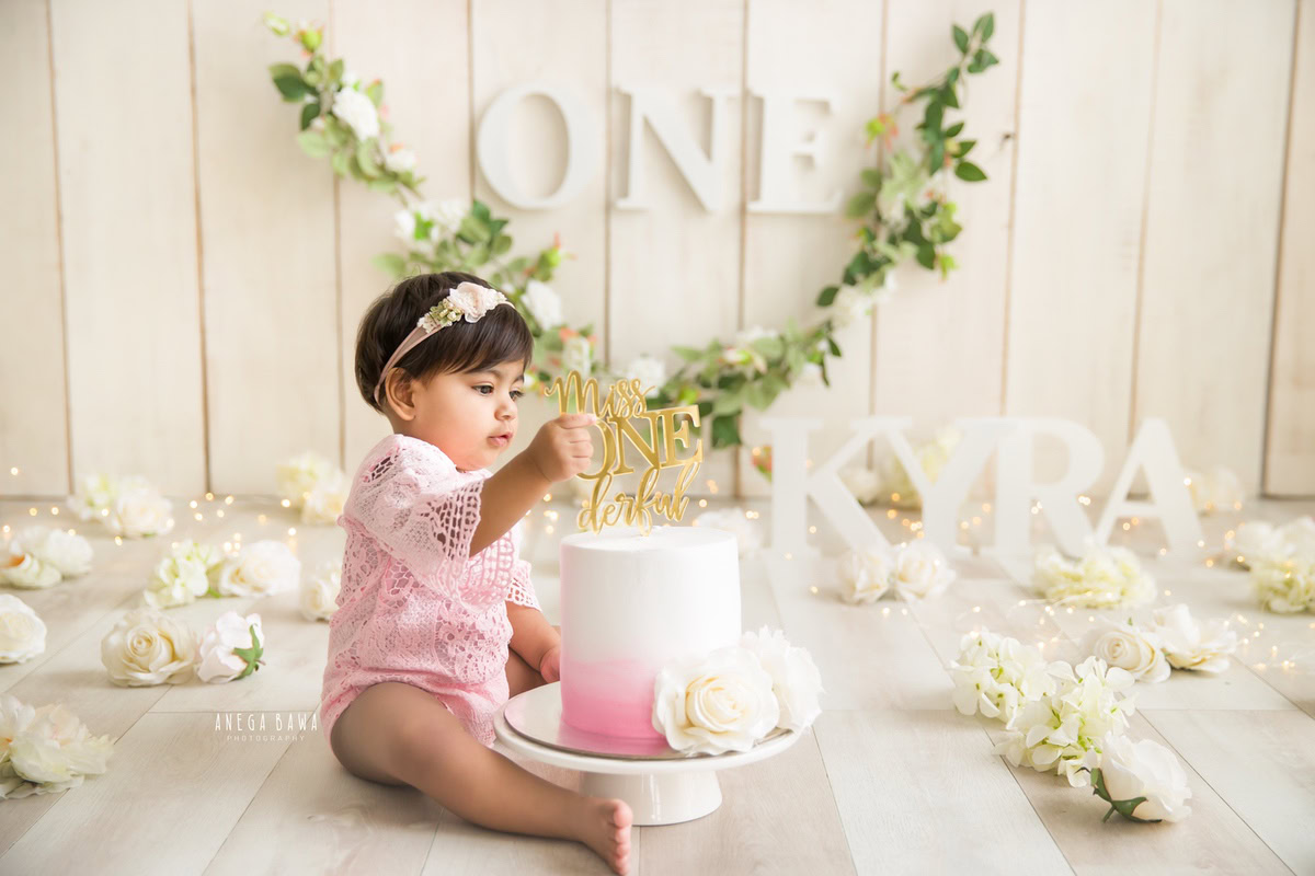 1 year old girl sitting on the floor with a cake smash setup, featuring white flowers, a floral fringe on the wall, and a name frame on the floor against a beige backdrop. Celebrating her first birthday. Cake Smash photo shoot in Delhi, Gurgaon by Anega Bawa Family Photographer Gurgaon (Delhi NCR).