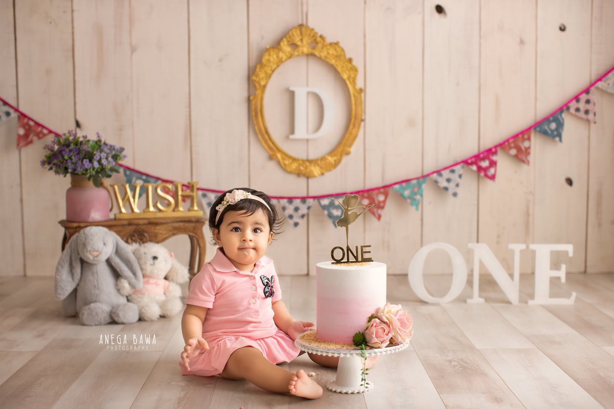 1 year old girl sitting with a cake smash setup, featuring a beige backdrop, colorful fringe on the wall, and a teddy bear. Celebrating her first birthday. Cake Smash photo shoot in Delhi, Gurgaon by Anega Bawa Family Photographer Gurgaon (Delhi NCR).