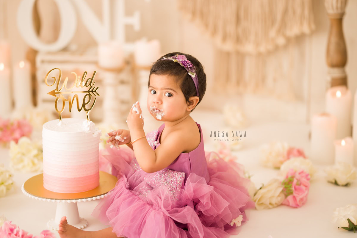 1year old girl sitting with a cake smash setup, featuring a candle stand and pink and white flowers, against a beige backdrop, captured by Anega Bawa during a Cake Smash photoshoot in Delhi, Gurgaon