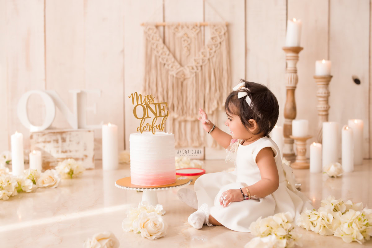 1 year old girl sitting with cake smash a candle stand and white flowers against a beige backdrop. Captured during a charming first birthday photoshoot in Delhi Gurgaon by the renowned family photographer Anega Bawa.