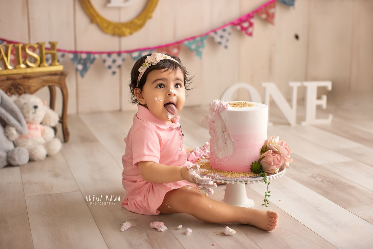 1 year old girl sitting with Cake Smash, featuring a colorful fringe on the wall, a cuddly teddy bear, and a beige backdrop, first birthday photography in Delhi, Gurgaon. Captured by Anega Bawa family photographer Gurgaon (Delhi NCR)