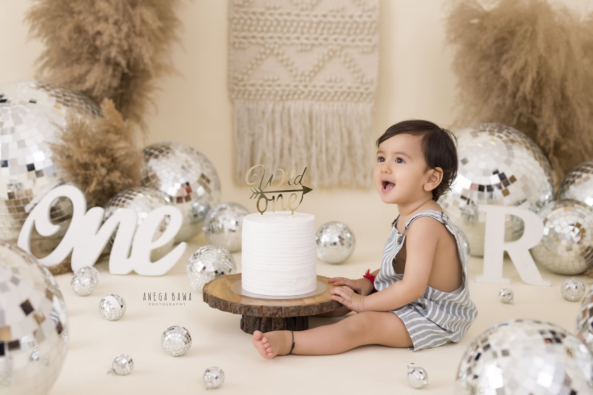 1year old girl sitting with a cake smash setup, featuring disco balls, bushes, and a net, against a beige backdrop, captured by Anega Bawa during a Cake Smash photoshoot in Delhi, Gurgaon