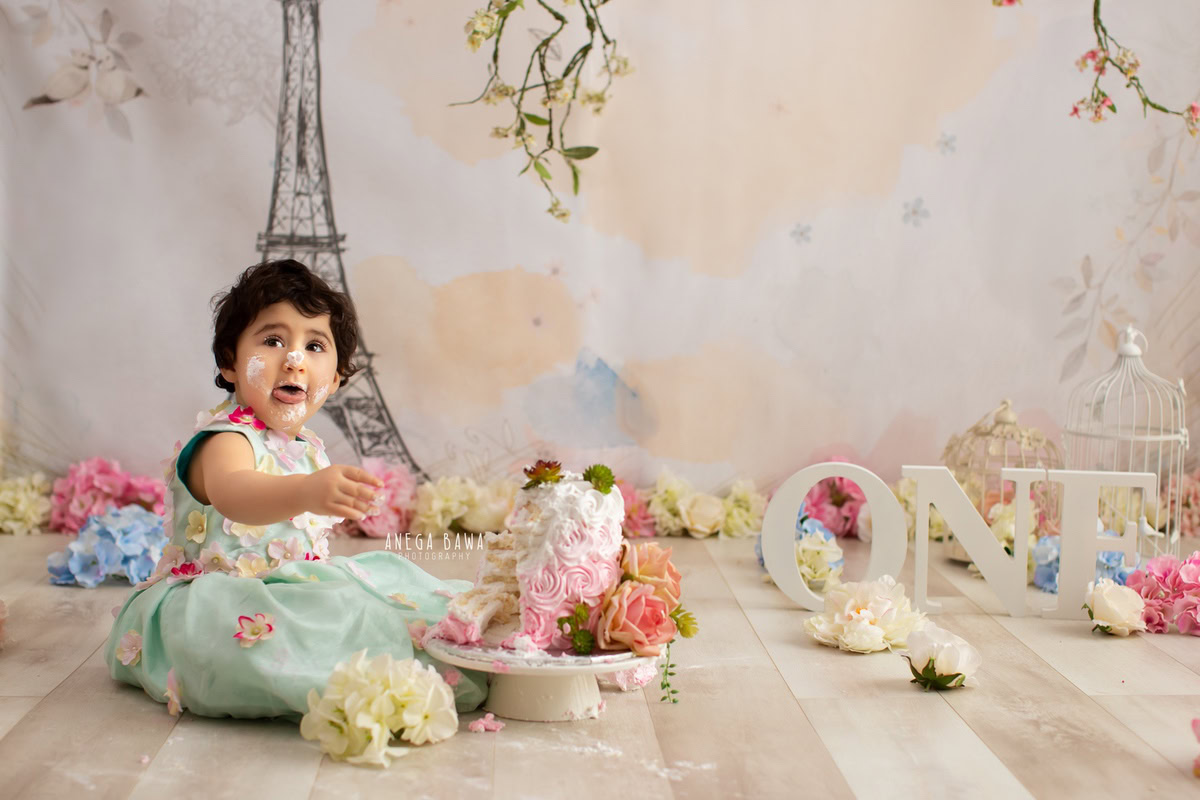 1 year old girl sitting with a cake smash setup, featuring flowers and an Eiffel Tower backdrop. Celebrating her first birthday. Cake Smash photo shoot in Delhi, Gurgaon by Anega Bawa Family Photographer Gurgaon (Delhi NCR).