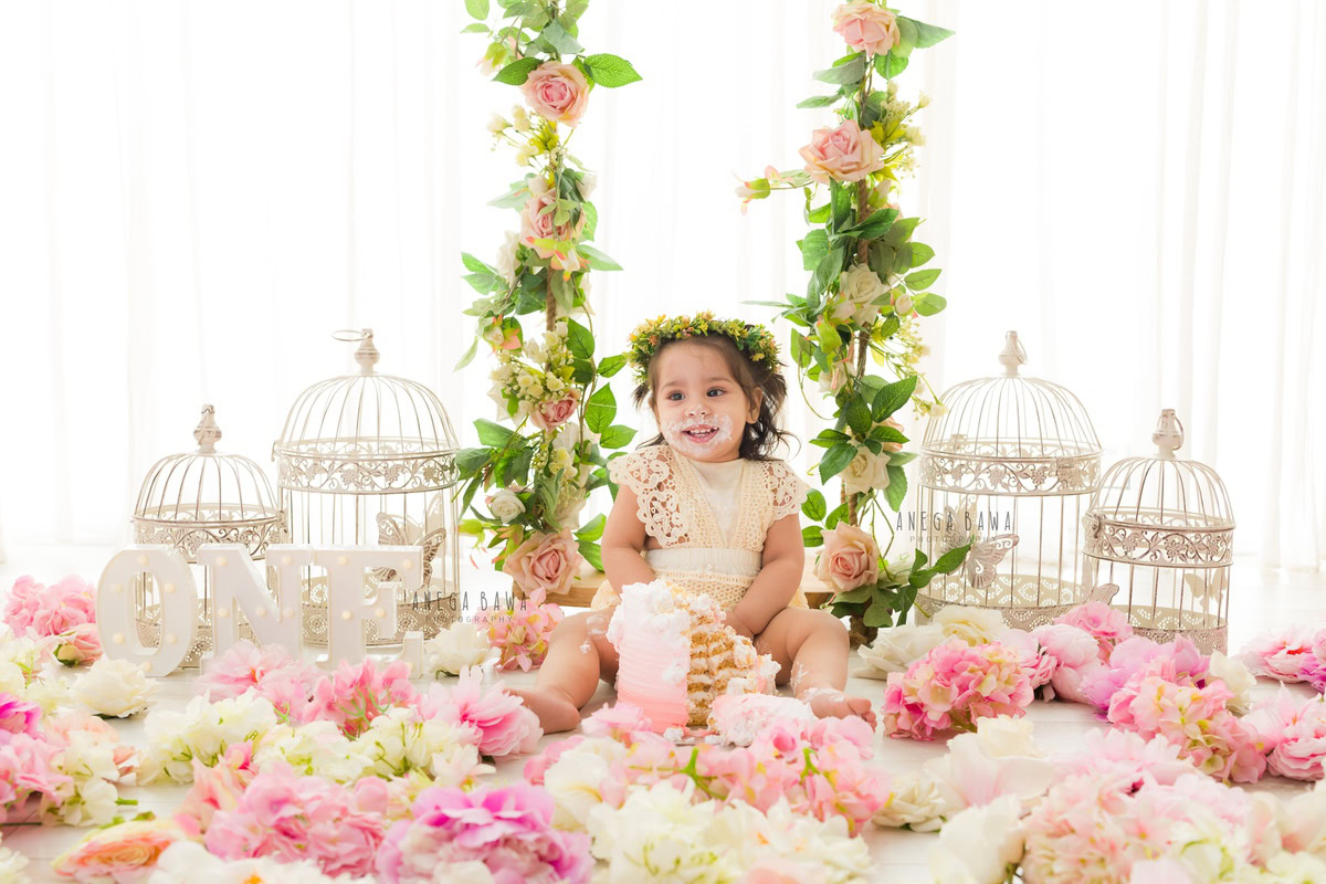 1 year old girl sitting with cake smash flowers on the floor white castles and a white backdrop. Captured during a delightful first birthday photoshoot in Delhi Gurgaon by the renowned family photographer Anega Bawa.