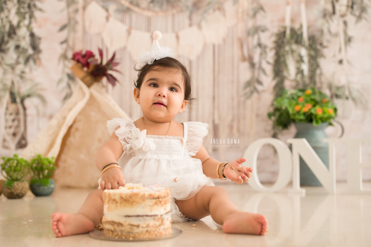1 year old girl sitting with cake smash a green vase and a peach and white backdrop. Captured during a charming first birthday photoshoot in Delhi Gurgaon by the renowned family photographer Anega Bawa.