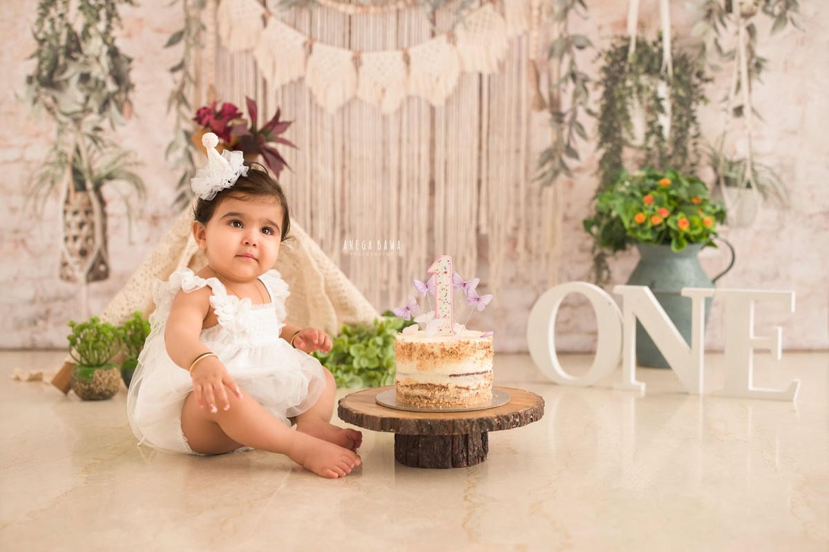 1 year old girl sitting with Cake Smash, vibrant peach and white backdrop, elegant green vase, first birthday photography in Delhi, Gurgaon. Captured by Anega Bawa family photographer Gurgaon (Delhi NCR)