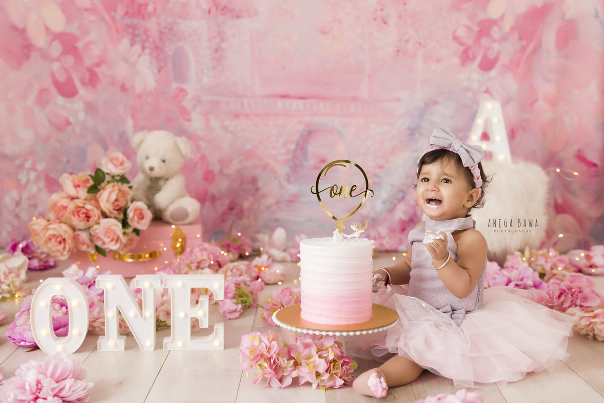 1 year old girl sitting with a cake smash setup, featuring a pink backdrop, pink and peach flowers, and a teddy bear. Celebrating her first birthday. Cake Smash photo shoot in Delhi, Gurgaon by Anega Bawa Family Photographer Gurgaon (Delhi NCR).