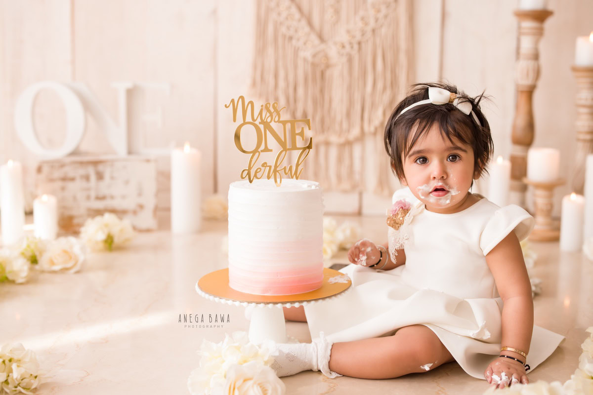 1 year old girl sitting with Cake Smash, white candles, beige backdrop, and a charming candle stand, first birthday photoshoot in Delhi, Gurgaon. Captured by Anega Bawa family photographer Gurgaon (Delhi NCR)