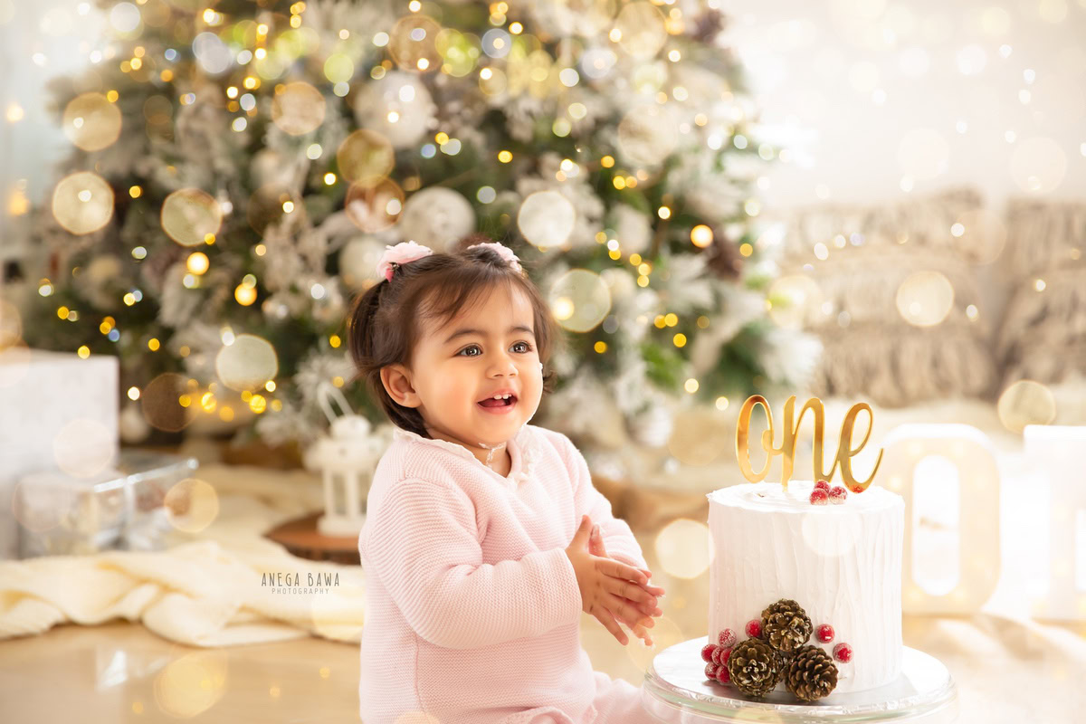 1 year old girl sitting with a cake smash setup, featuring a Christmas tree and a white backdrop. Celebrating her first birthday. Cake Smash photo shoot in Delhi, Gurgaon by Anega Bawa Family Photographer Gurgaon (Delhi NCR).