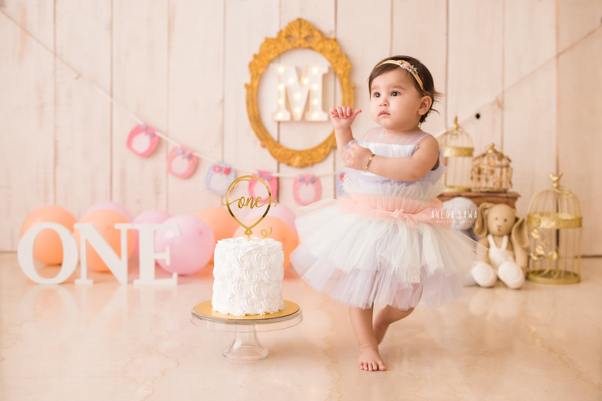 1 year old girl standing with a cake smash setup, featuring golden castles, teddy bears, pink and peach balloons, and a beige backdrop. Celebrating her first birthday. Cake Smash photo shoot in Delhi, Gurgaon by Anega Bawa Family Photographer Gurgaon (Delhi NCR).