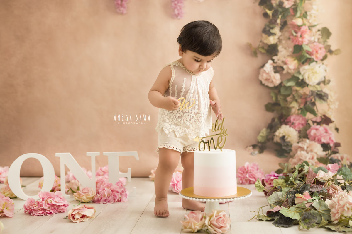 1 year old girl standing with a cake smash setup, featuring a peach backdrop, floral fringe on the wall, and pink flowers on the floor. Celebrating her first birthday. Cake Smash photo shoot in Delhi, Gurgaon by Anega Bawa Family Photographer Gurgaon (Delhi NCR).