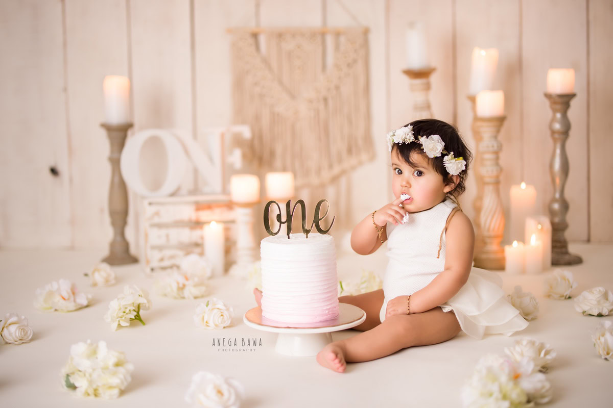 1 year old girl with a tiara band, a candle stand, and flowers on the floor, set against a beige backdrop. Celebrating her first birthday. Cake Smash photo shoot in Delhi, Gurgaon by Anega Bawa Family Photographer Gurgaon (Delhi NCR).
