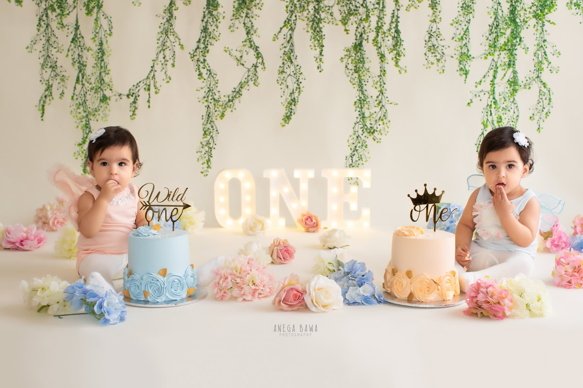1 year old twins with a cake smash setup, featuring leafy fringes on the wall, multicolored flowers, and a white backdrop. Celebrating their first birthday. Cake Smash photo shoot in Delhi, Gurgaon by Anega Bawa Family Photographer Gurgaon (Delhi NCR).