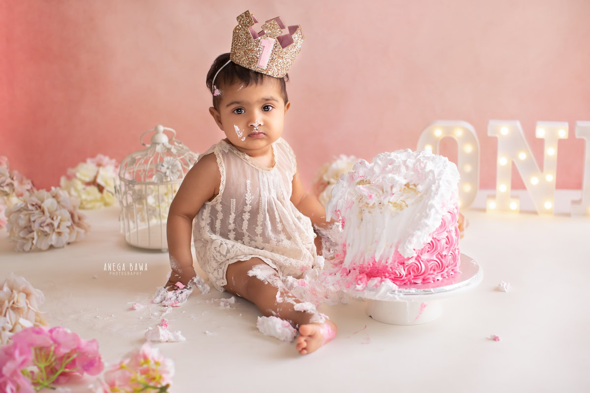 1 year old girl wearing a crown and playing with cake smash against a pink backdrop with a castle. Captured during a charming first birthday photo shoot in Delhi Gurgaon by the renowned family photographer Anega Bawa.